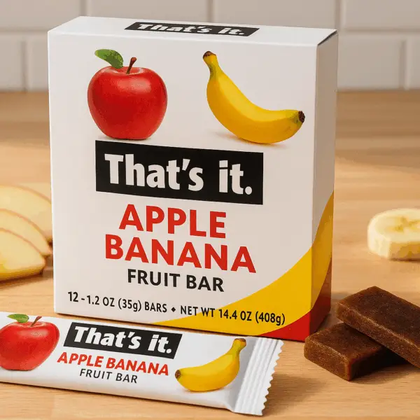 A box of That's it. Apple Banana Fruit Bars on a wooden countertop, with one unwrapped bar, apple slices, and banana pieces beside it.