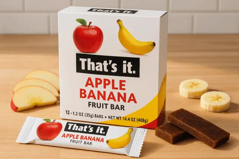 A box of That's it. Apple Banana Fruit Bars on a wooden countertop, with one unwrapped bar, apple slices, and banana pieces beside it.