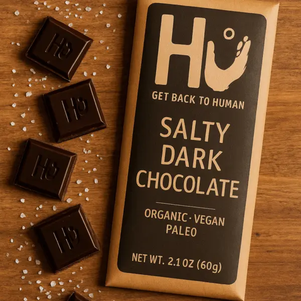 A Hu Salty Dark Chocolate Bar on a wooden surface, with broken chocolate squares and sea salt scattered around the packaging.